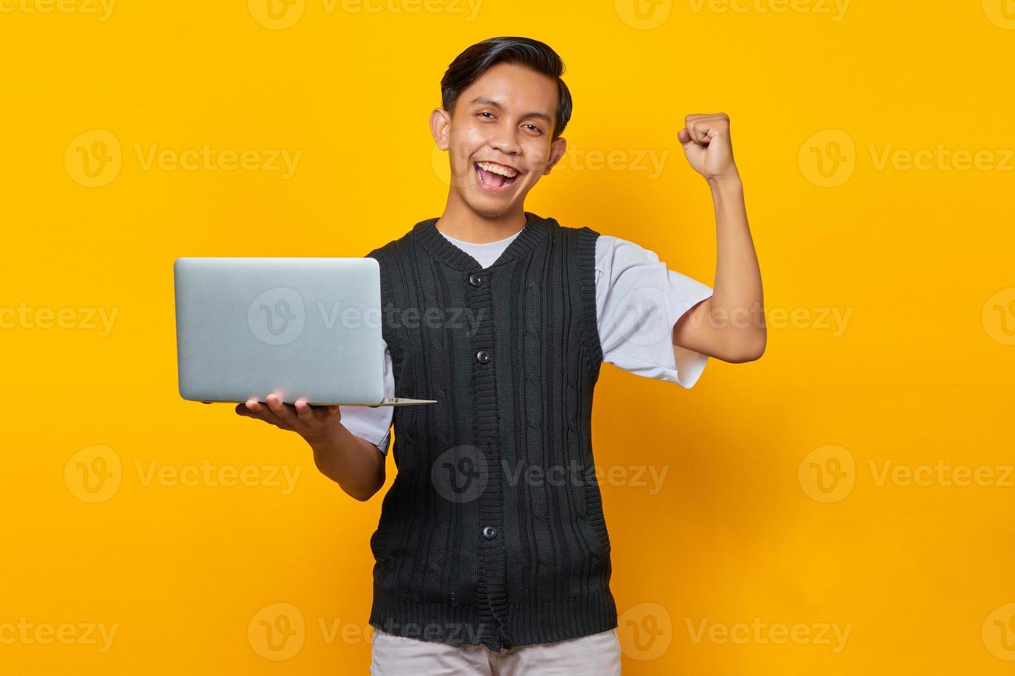 Homem asiático animado segurando um laptop e comemorando a vitória, sorrindo sobre fundo amarelo foto