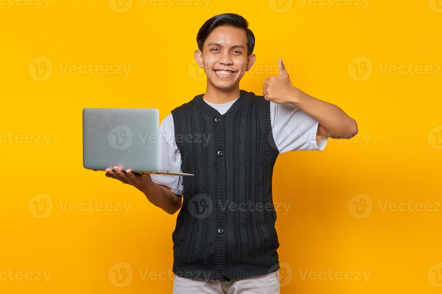 bonito homem asiático segurando laptop sorrindo e mostrando os polegares para cima sinal sobre fundo amarelo foto