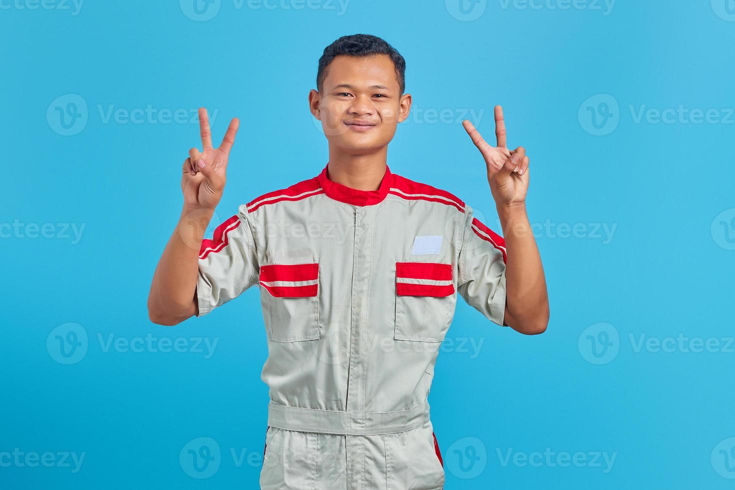 retrato de um jovem mecânico asiático sorridente, fazendo o sinal da paz com o dedo isolado no fundo azul foto