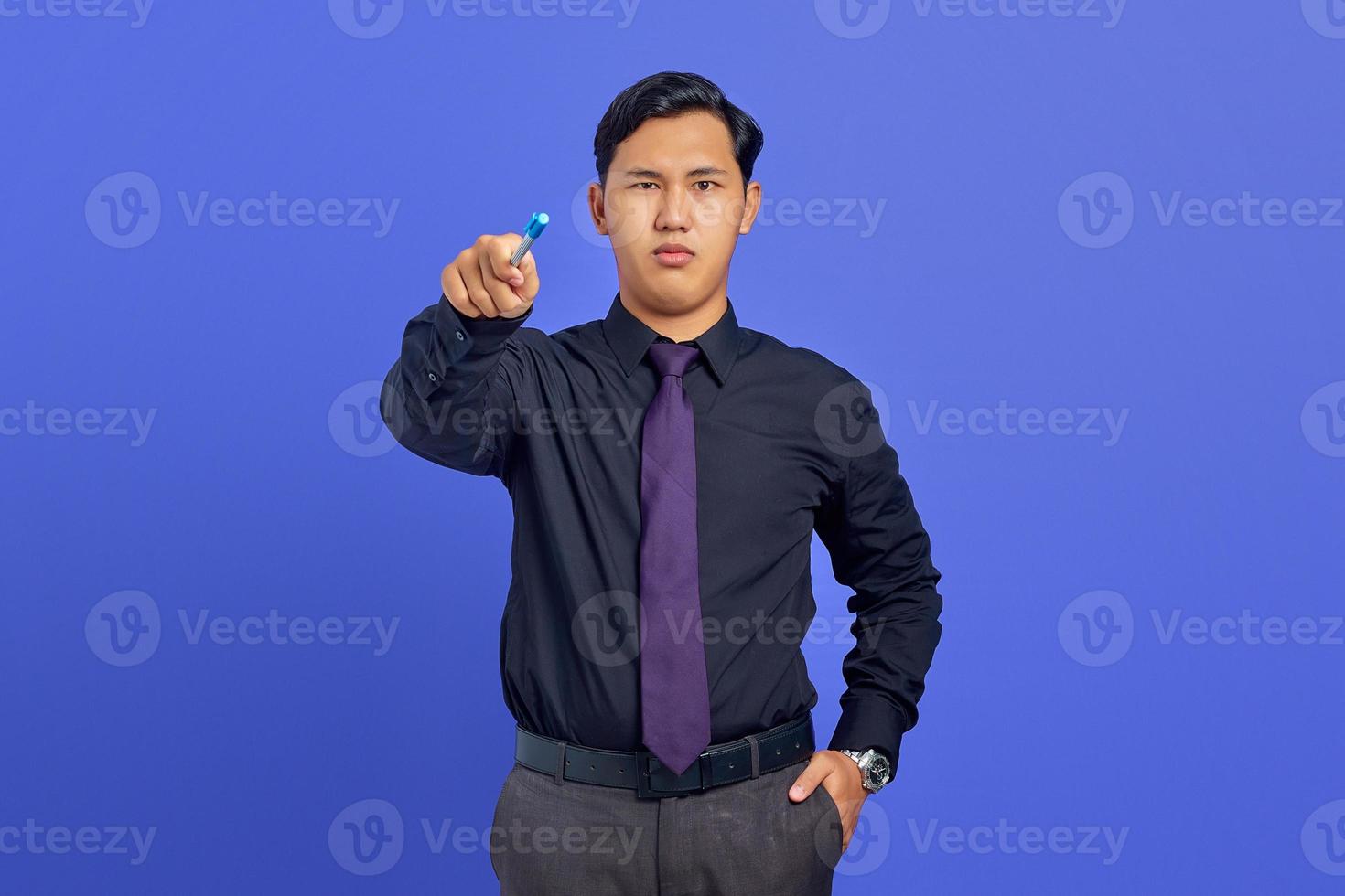 Belo jovem empresário sério segurando uma caneta e apontando para a câmera em fundo roxo foto