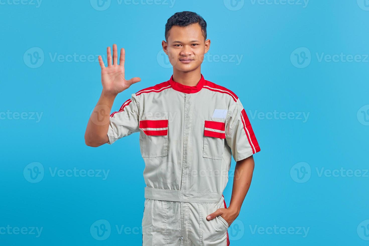 retrato de um jovem mecânico asiático sorridente, fazendo o sinal de número cinco com o dedo isolado no fundo azul foto