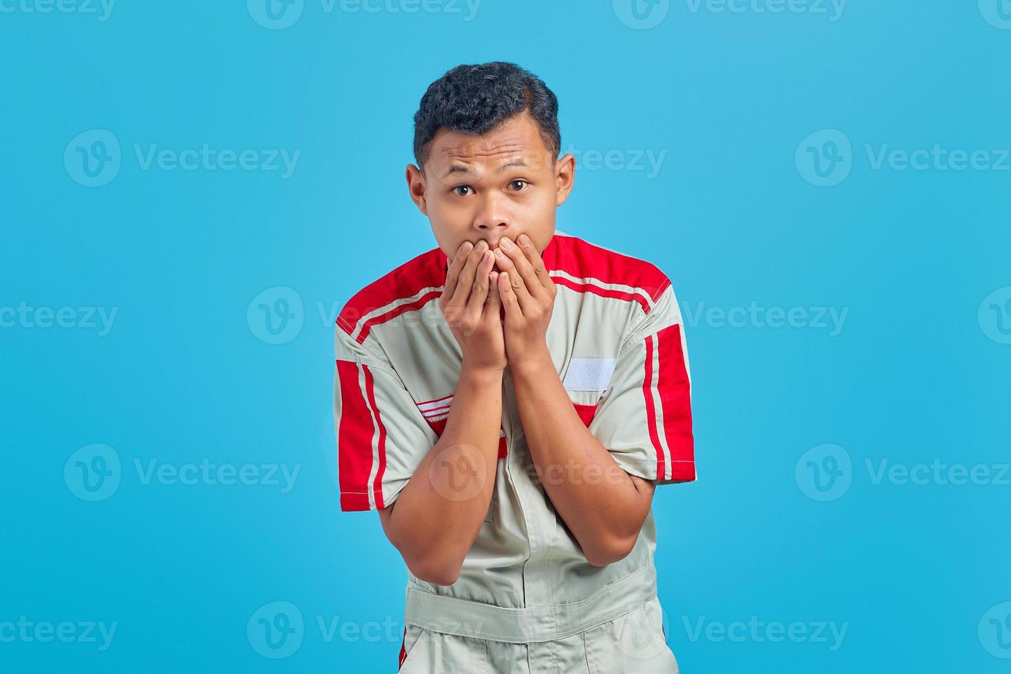 retrato de jovem mecânico asiático cobrindo a boca com as mãos com expressão de surpresa no fundo azul foto