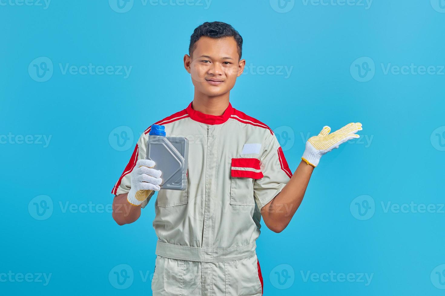 retrato de um jovem mecânico asiático sorridente, mostrando a garrafa de plástico de óleo de máquina e apontando para copiar o espaço com a palma da mão sobre o fundo azul foto