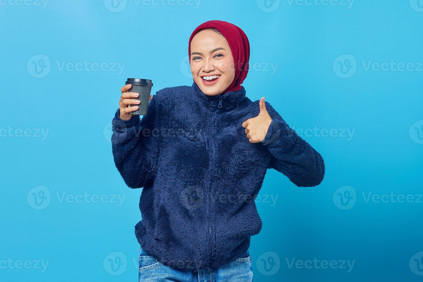 alegre linda jovem segurando a xícara de café e mostrando o gesto de polegar para cima sobre o fundo azul foto