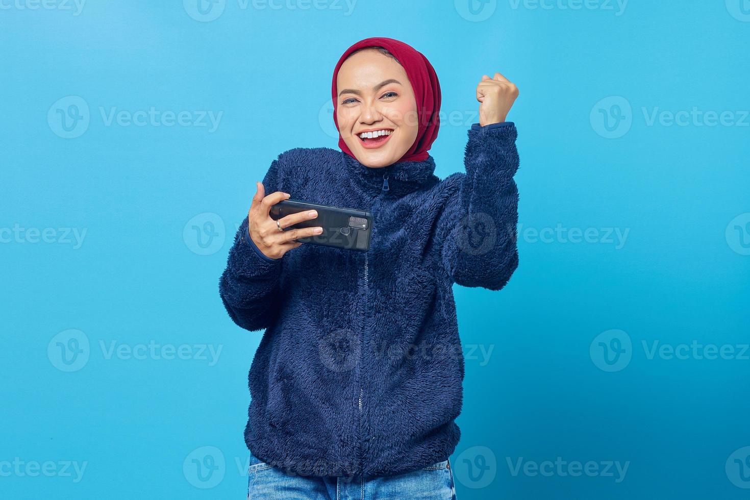 alegre mulher asiática jogando um jogo em um telefone celular enquanto celebra a vitória sobre fundo azul foto