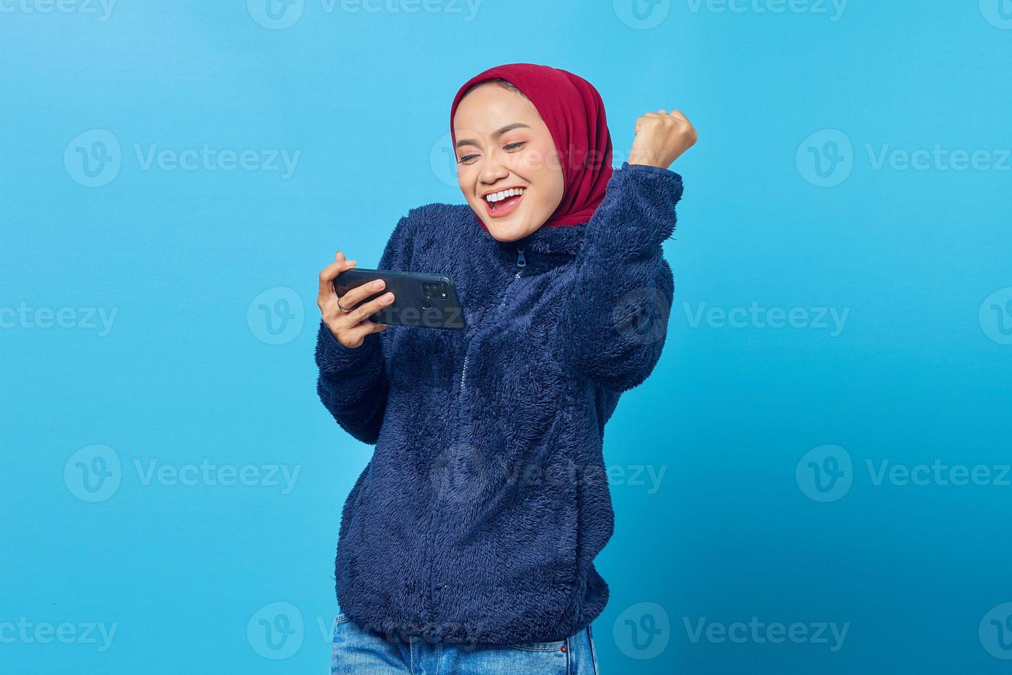 Mulher asiática animada jogando um jogo em um telefone celular enquanto celebra a vitória sobre fundo azul foto