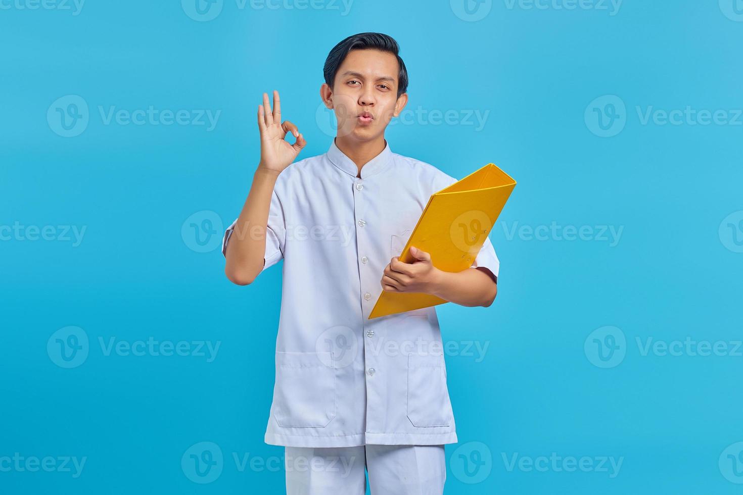 retrato de alegre enfermeira masculina asiática segurando uma pasta amarela e fazendo gesto de ok sobre fundo azul foto