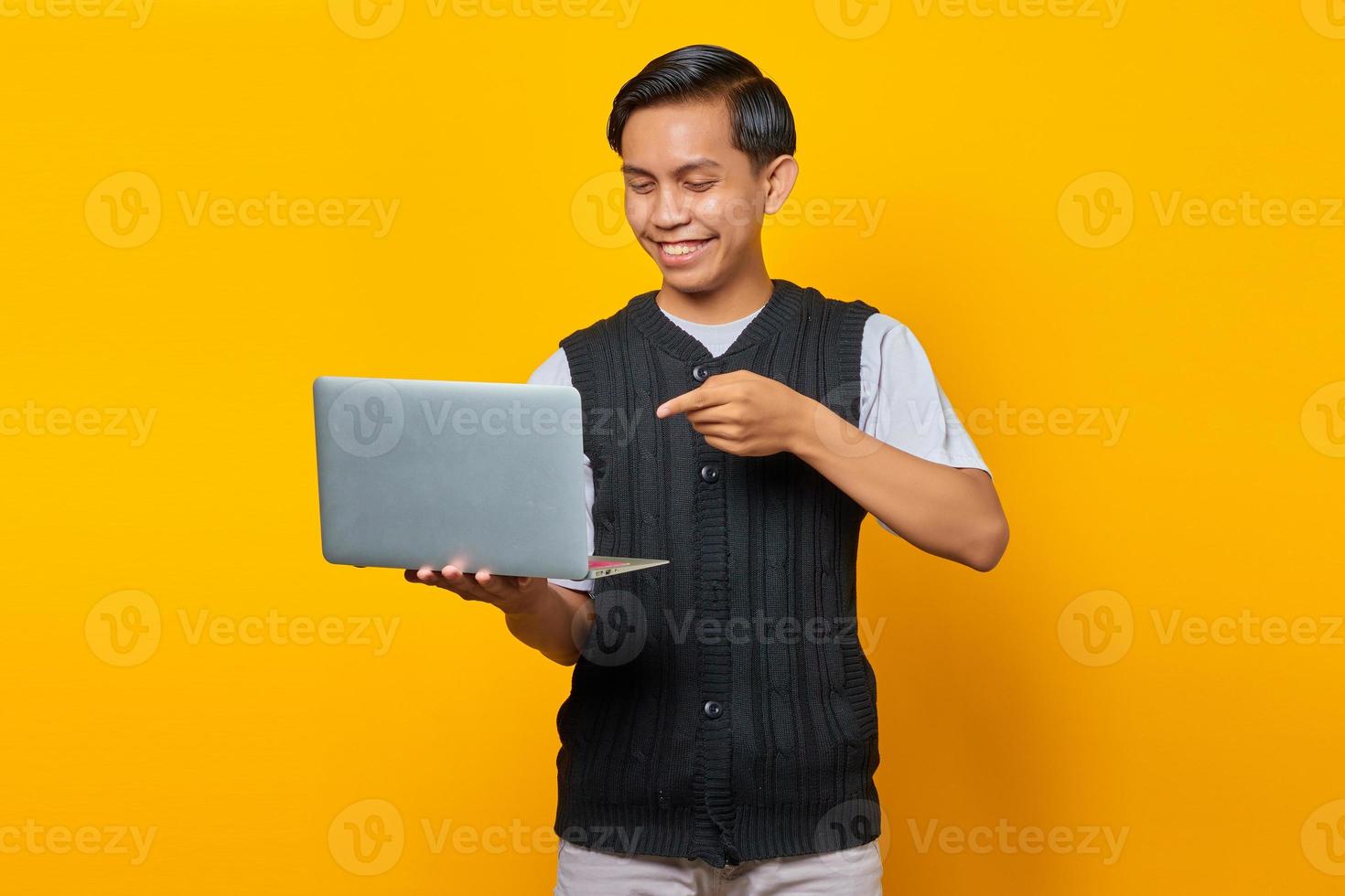 jovem bonito alegre apontando para laptop sobre fundo amarelo foto