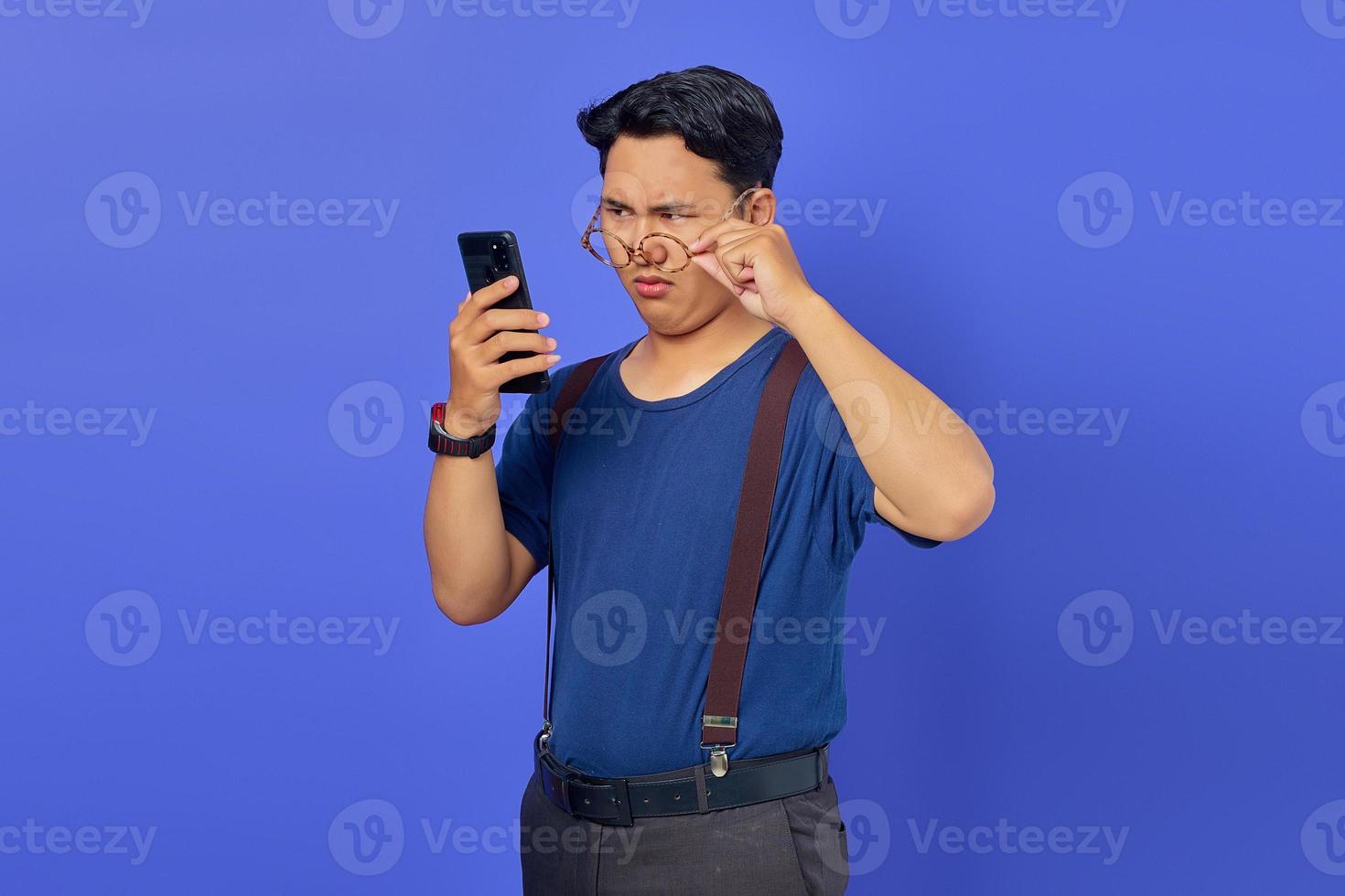 jovem asiático chocado olhando para a tela do smartphone e tirando os óculos no fundo roxo foto