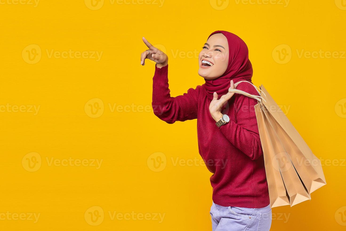 jovem mulher asiática feliz segurando uma sacola de compras e apontando para um espaço vazio no fundo amarelo foto