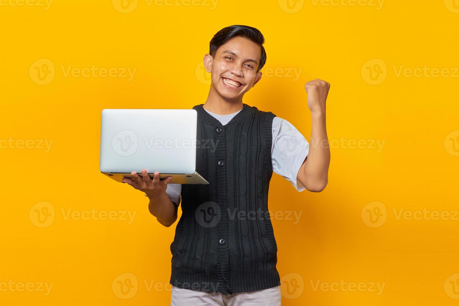 Homem asiático animado segurando um laptop e comemorando a vitória, sorrindo sobre fundo amarelo foto