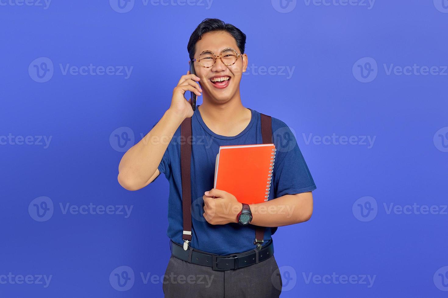 alegre jovem estudante falando no smartphone enquanto segura o caderno no fundo roxo foto