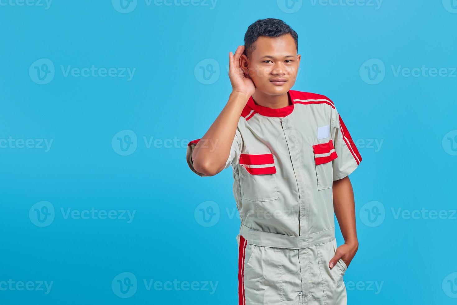 Um mecânico asiático sério tentando ouvir uma conversa secreta isolada sobre fundo azul foto