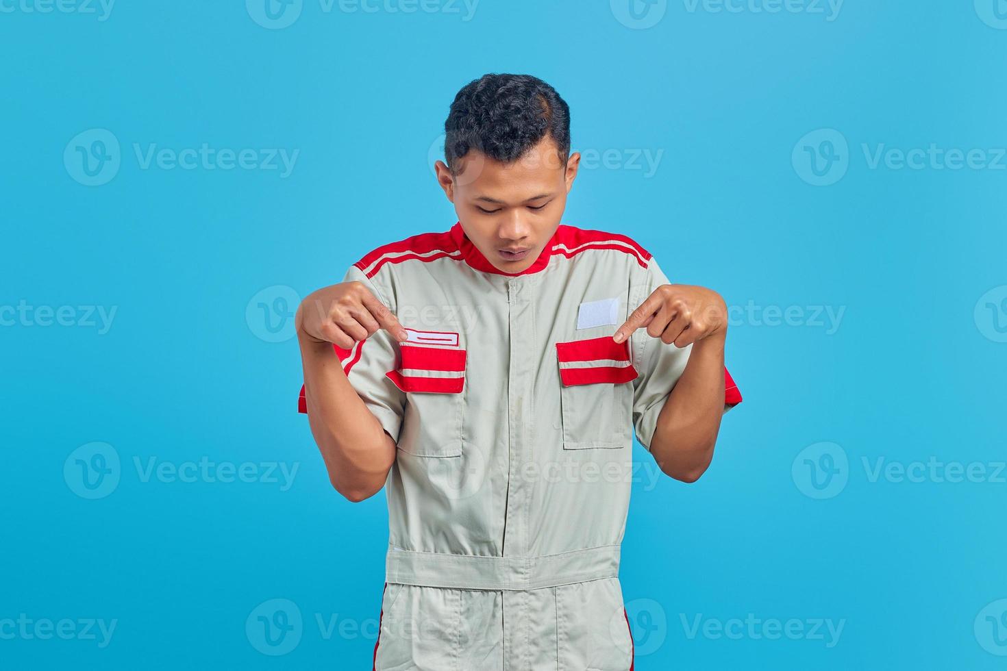 jovem mecânico chocado olhando para baixo e apontando para baixo com o dedo isolado no fundo azul foto