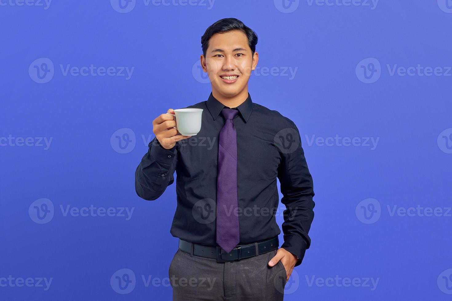 belo jovem empresário sorrindo para a câmera com as mãos no bolso e segurando uma caneca no fundo roxo foto