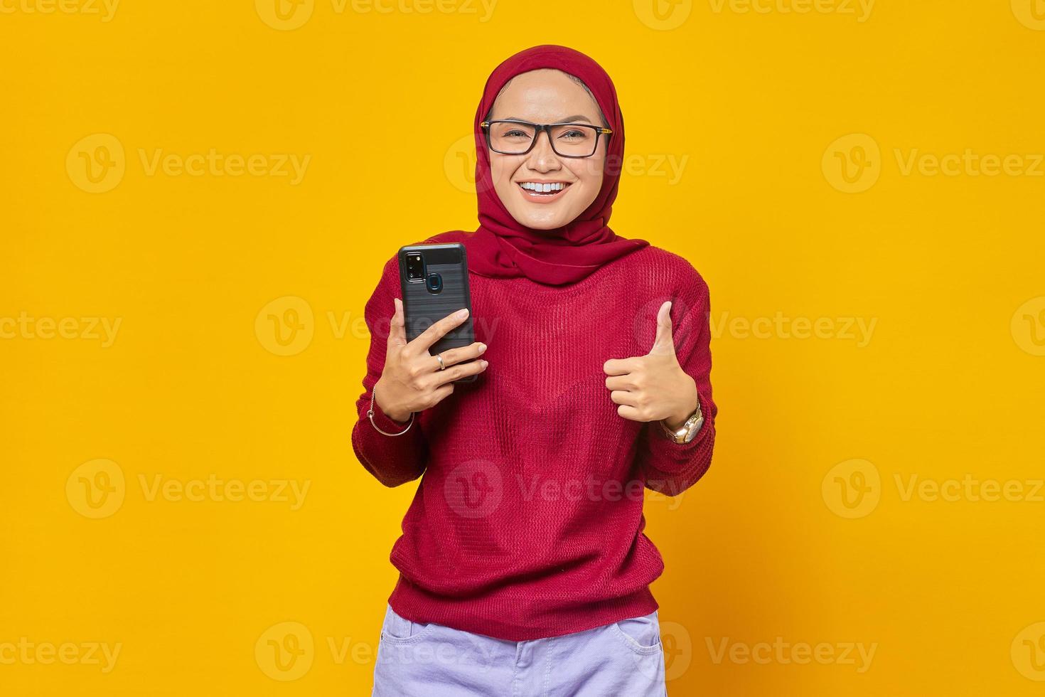 retrato de uma jovem asiática alegre segurando um telefone celular e mostrando os polegares para cima sinal isolado em fundo amarelo foto