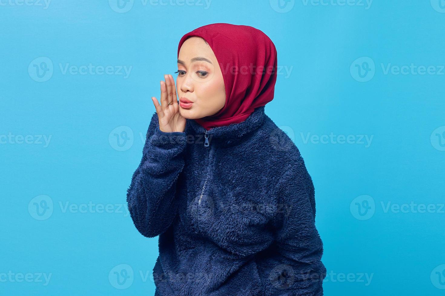 retrato de uma linda mulher asiática mantém a mão perto da boca e sussurra um segredo sobre fundo azul. foto