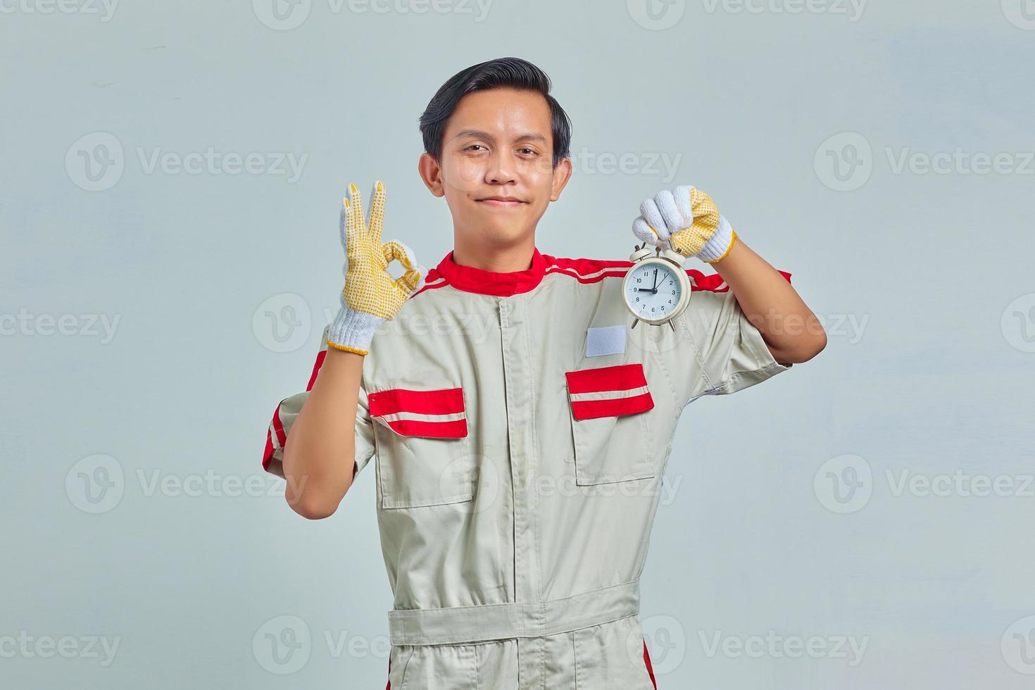 retrato de homem bonito alegre, vestindo uniforme de mecânico, segurando o relógio despertador e mostrando sinal de ok no fundo cinza foto