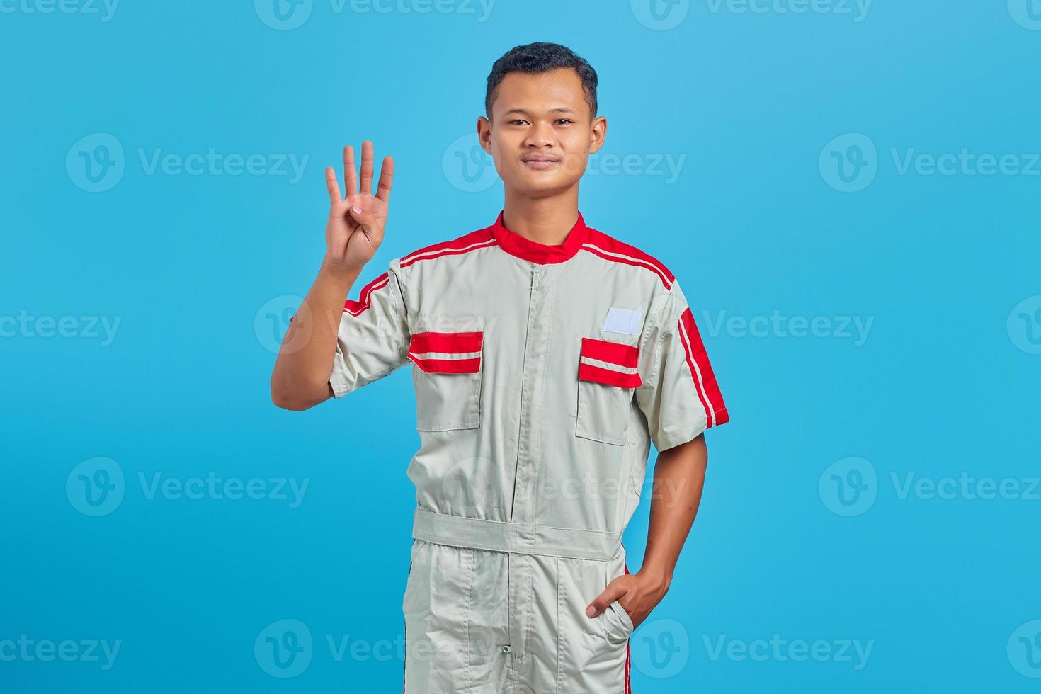 retrato de um jovem mecânico asiático sorridente, fazendo o sinal de número quatro com o dedo isolado no fundo azul foto