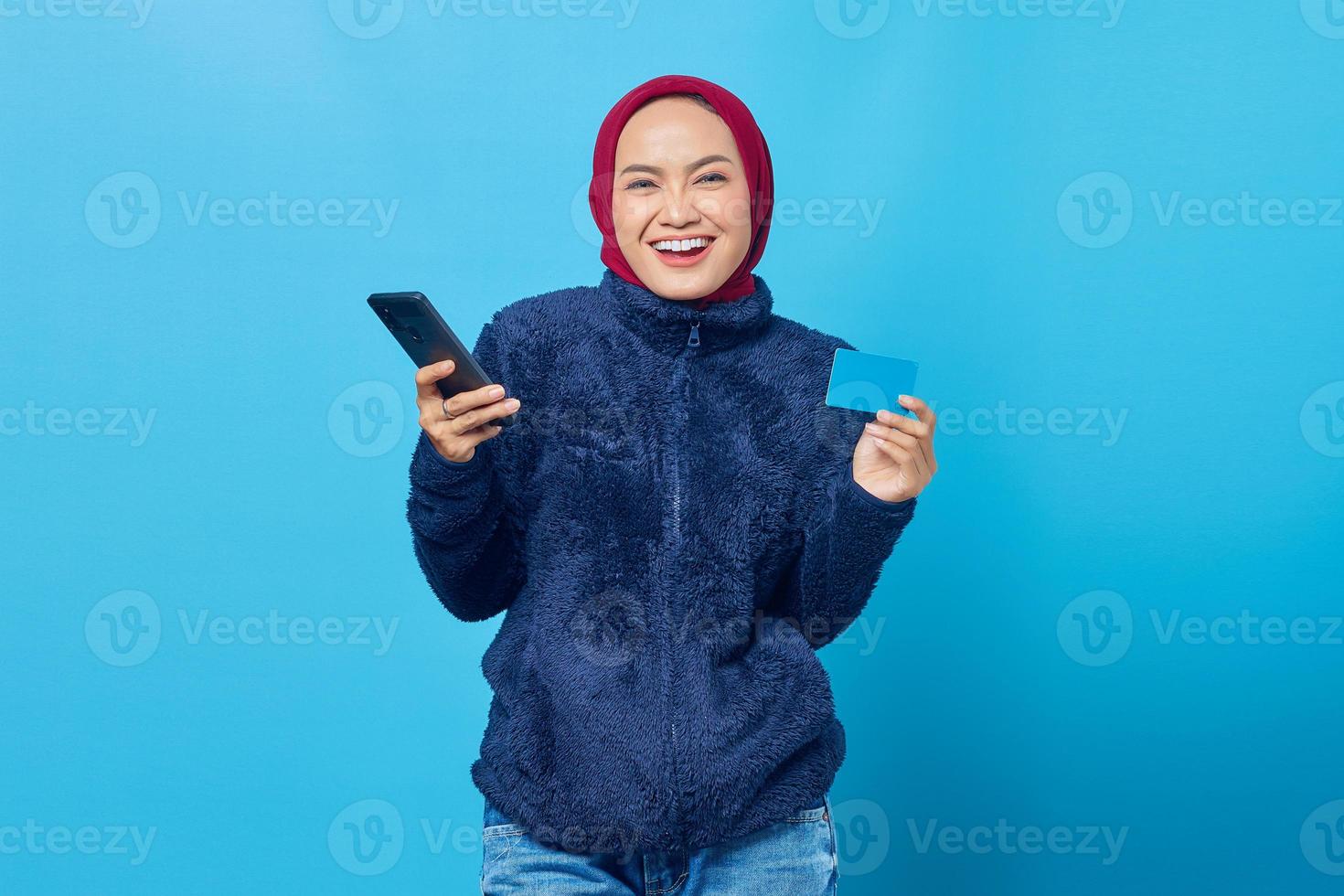 Retrato de uma jovem asiática sorridente, segurando um telefone celular e mostrando o cartão de crédito sobre fundo azul foto