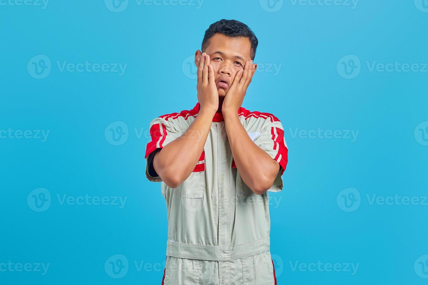 retrato de um jovem mecânico asiático tocando a bochecha com expressão entediada, isolado no fundo azul foto