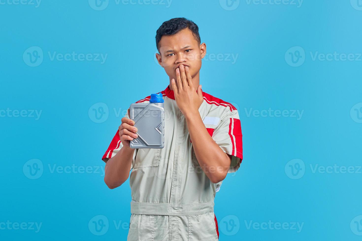 Retrato de jovem mecânico asiático chocado segurando uma garrafa de plástico de óleo de motor e cobrindo a boca com as mãos com fundo azul foto