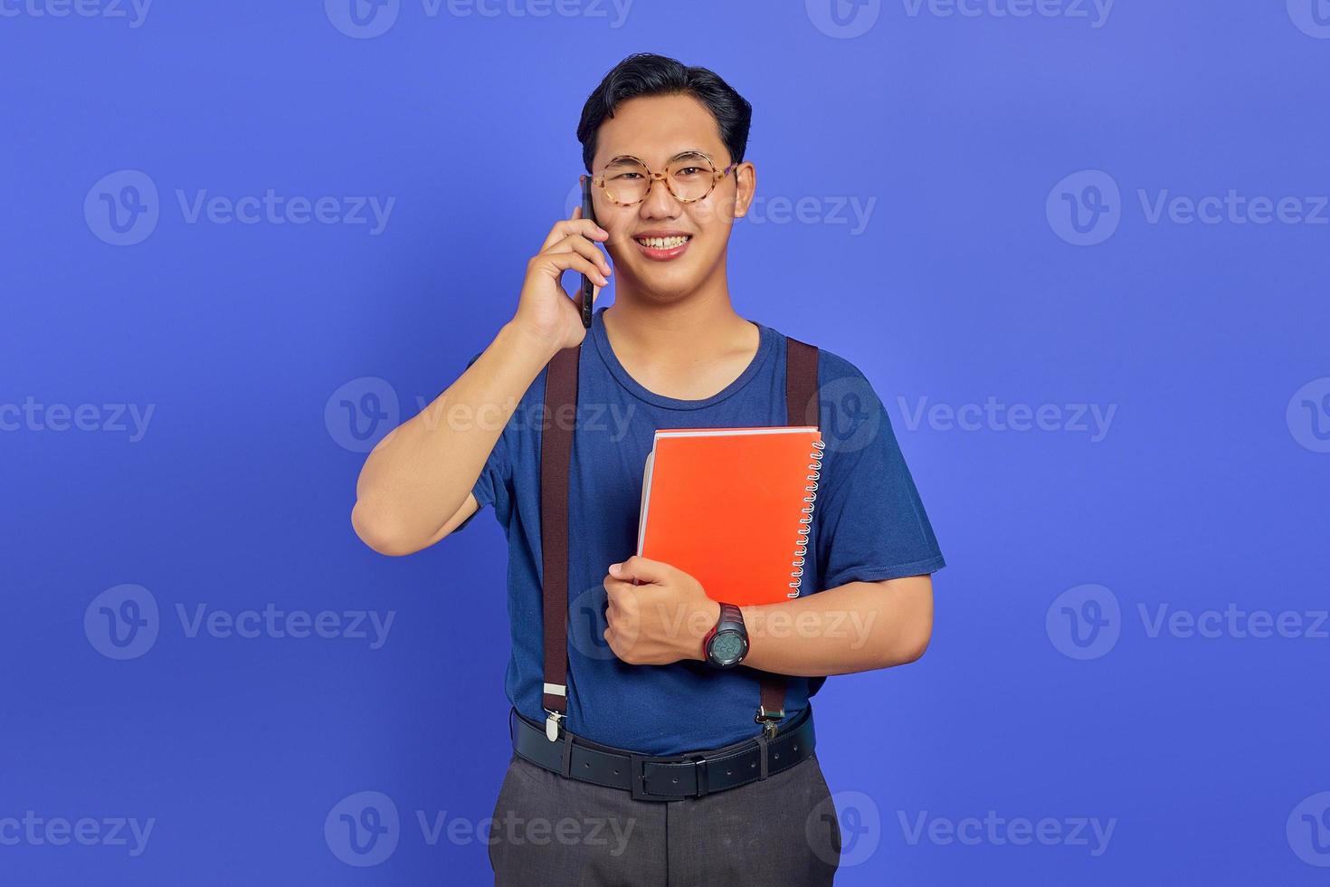 jovem e bonito estudante falando no smartphone enquanto segura o caderno no fundo roxo foto