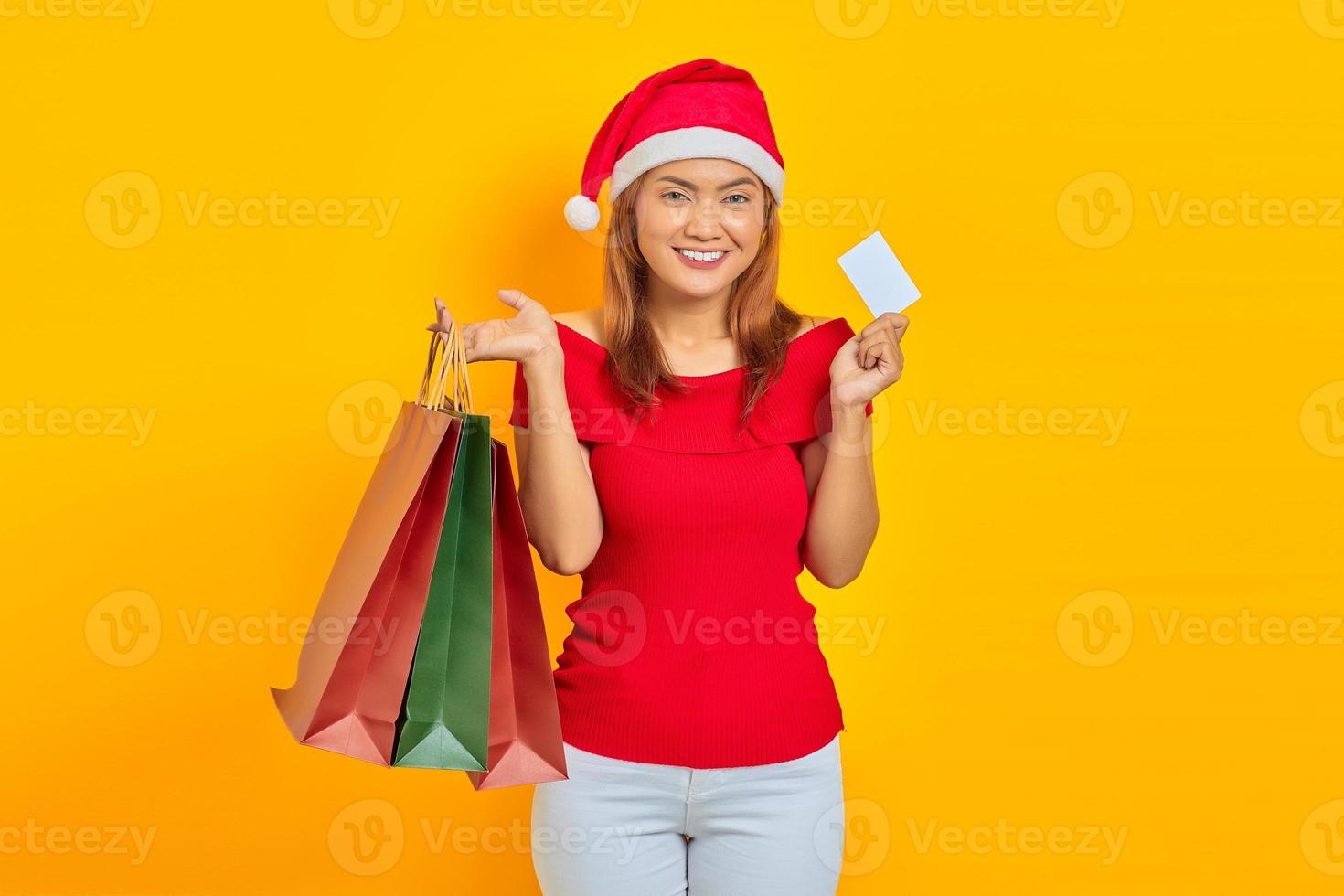 sorridente jovem mulher asiática com chapéu de Papai Noel segurando sacolas de compras e mostrando um cartão em branco sobre fundo amarelo foto