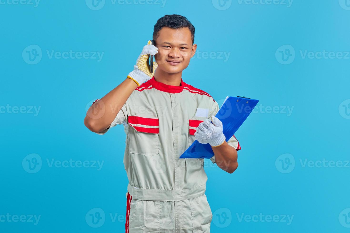 sorrindo, jovem mecânico asiático falando no celular e segurando a prancheta isolada sobre fundo azul foto