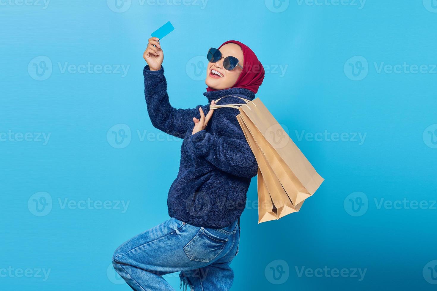 animada e alegre jovem mulher asiática segurando sacolas de compras e cartão de crédito sobre fundo azul foto