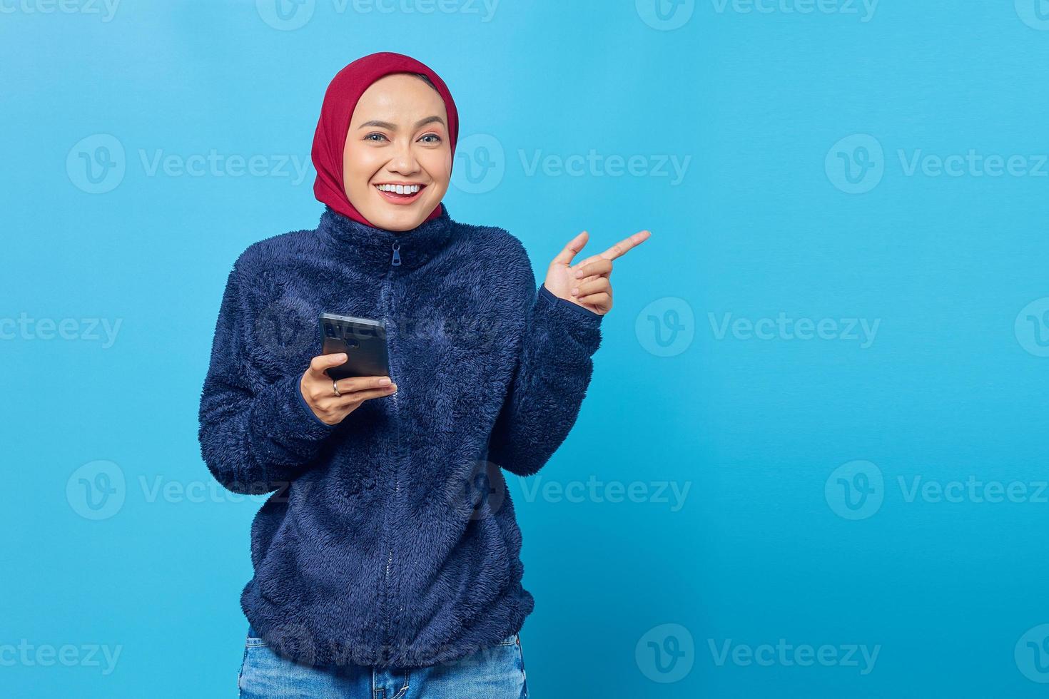 Mulher asiática sorridente segurando um telefone celular enquanto aponta o dedo para o espaço da cópia sobre fundo azul foto