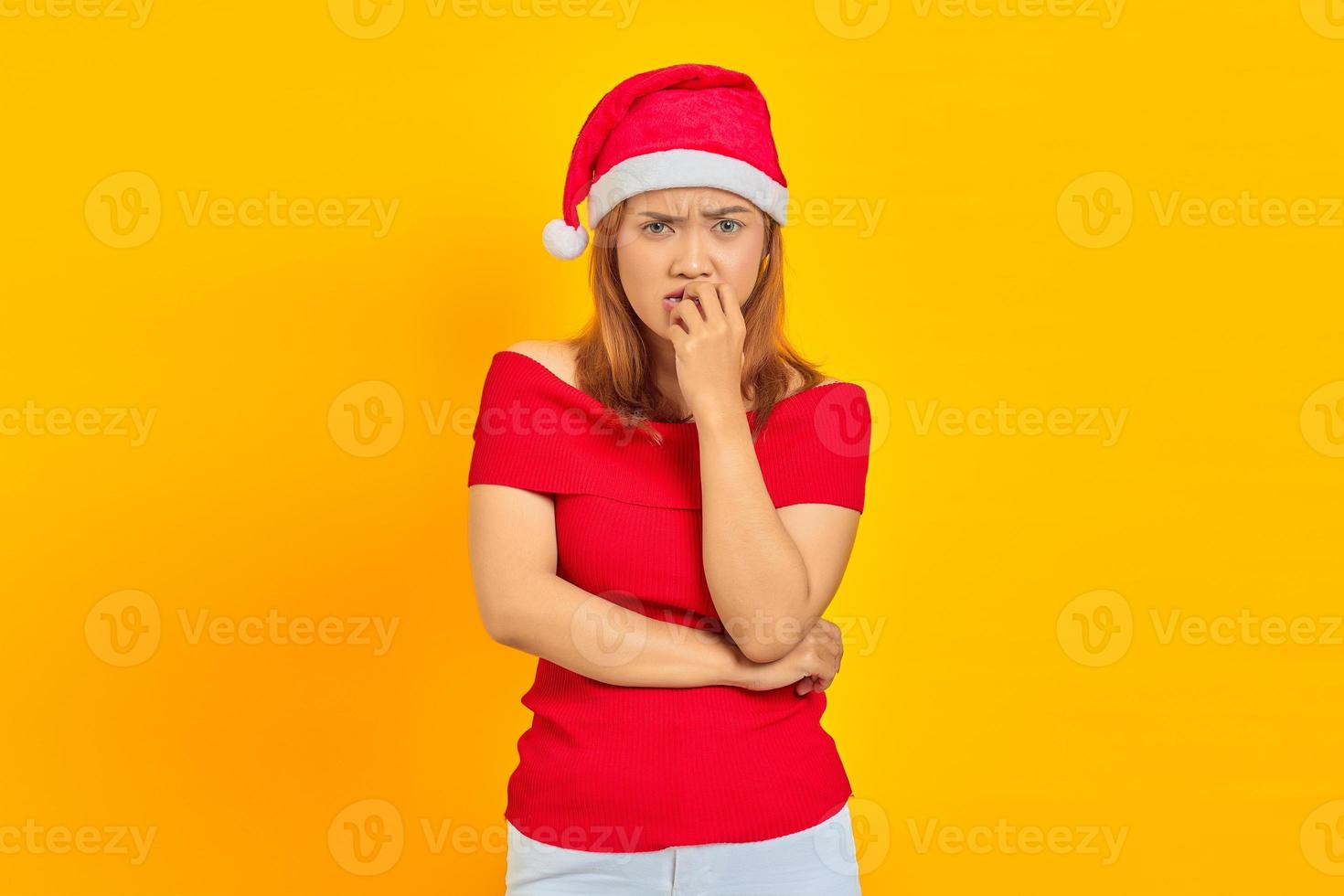 jovem asiática com chapéu de Natal, parecendo estressada e nervosa, com a mão na boca roendo as unhas foto