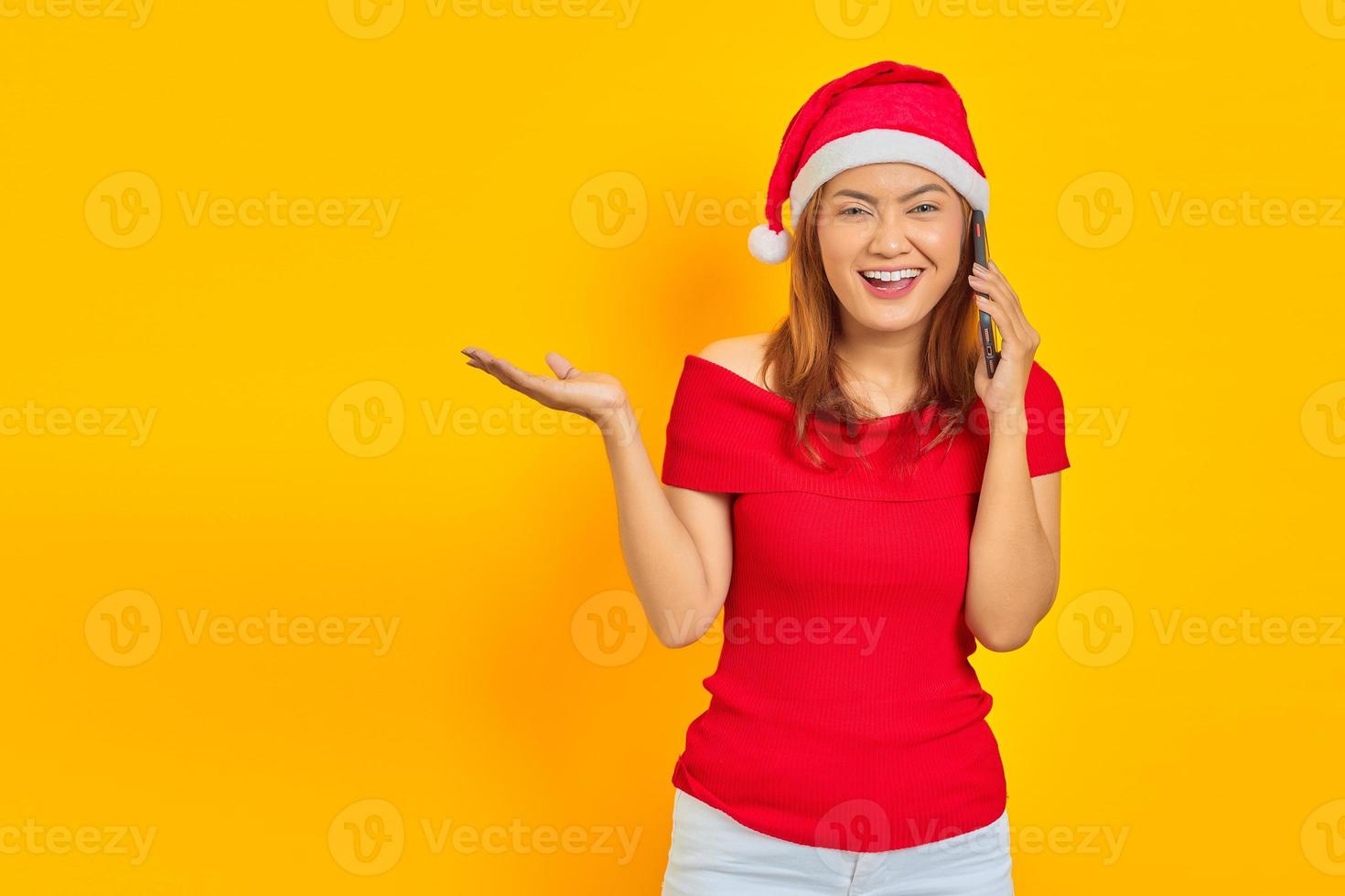 jovem asiática alegre usando chapéu de natal, falando no celular enquanto aponta para o lado foto