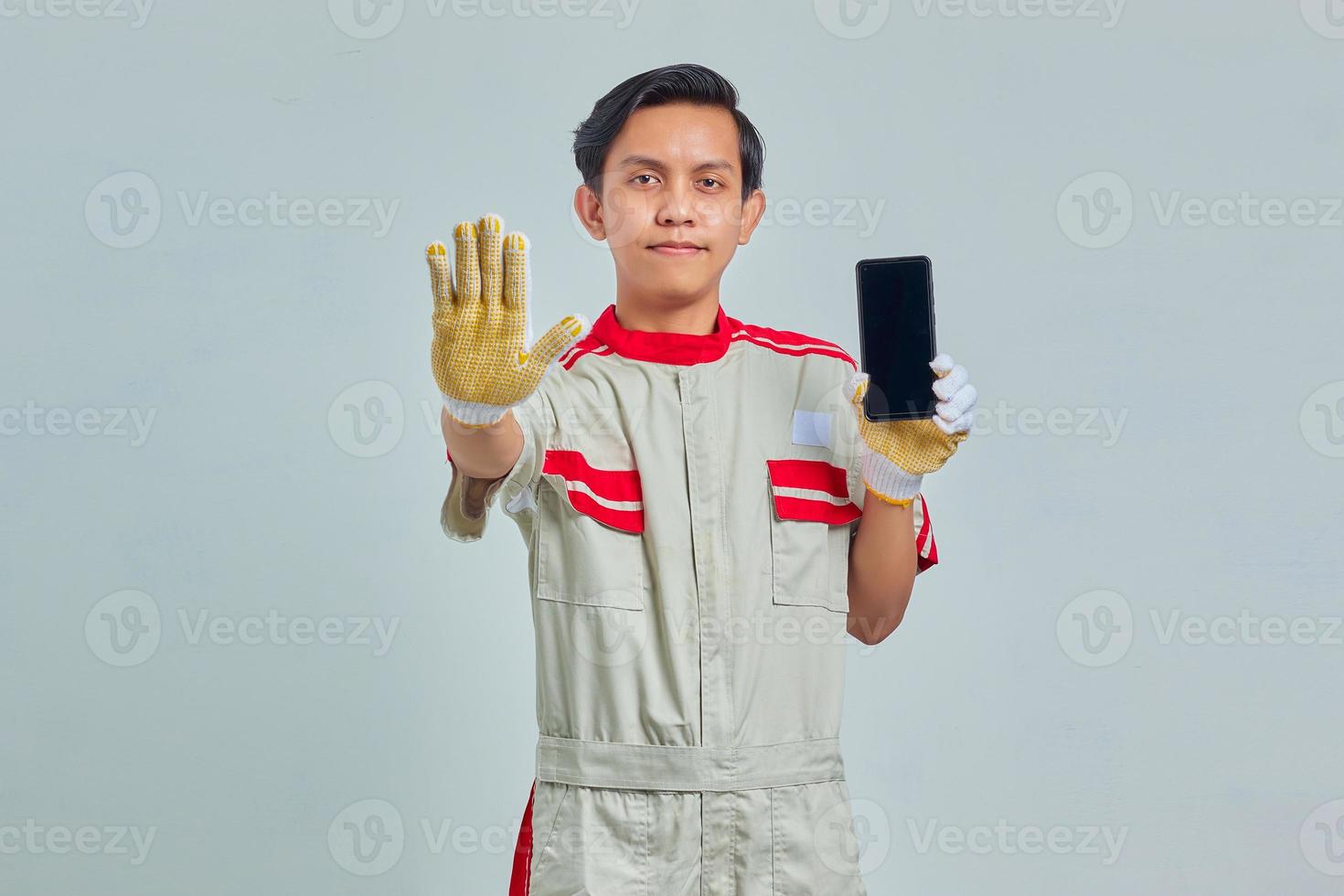 retrato de homem bonito usando uniforme mecânico, segurando o smartphone e fazendo gesto de parada com as palmas das mãos foto