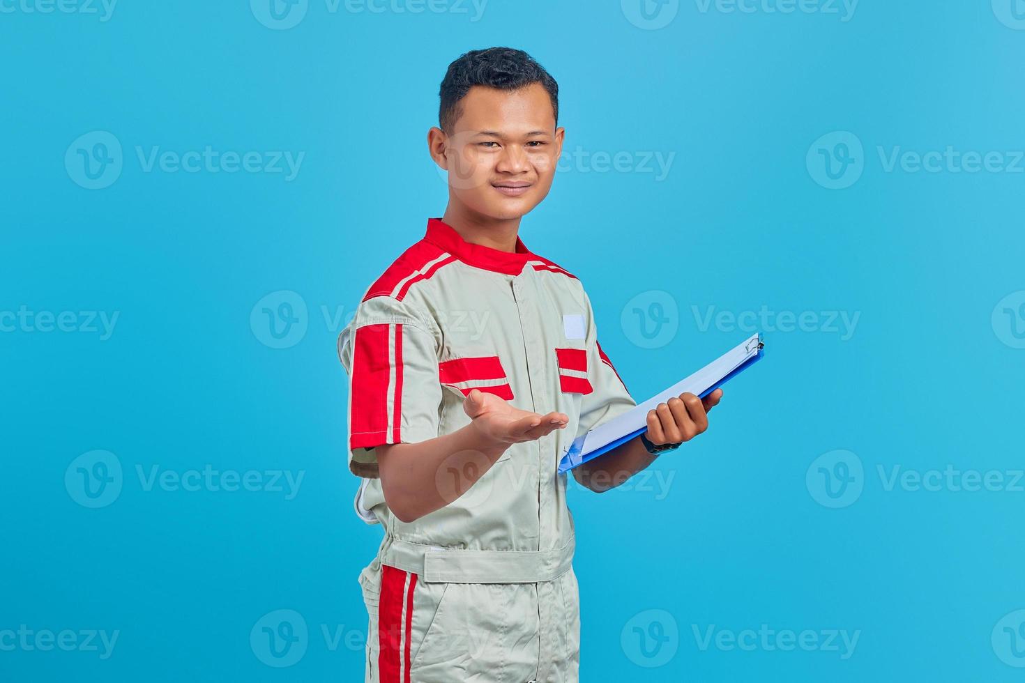 retrato de um jovem mecânico alegre segurando a prancheta e apontando com a palma da mão para a câmera isolada sobre fundo azul foto