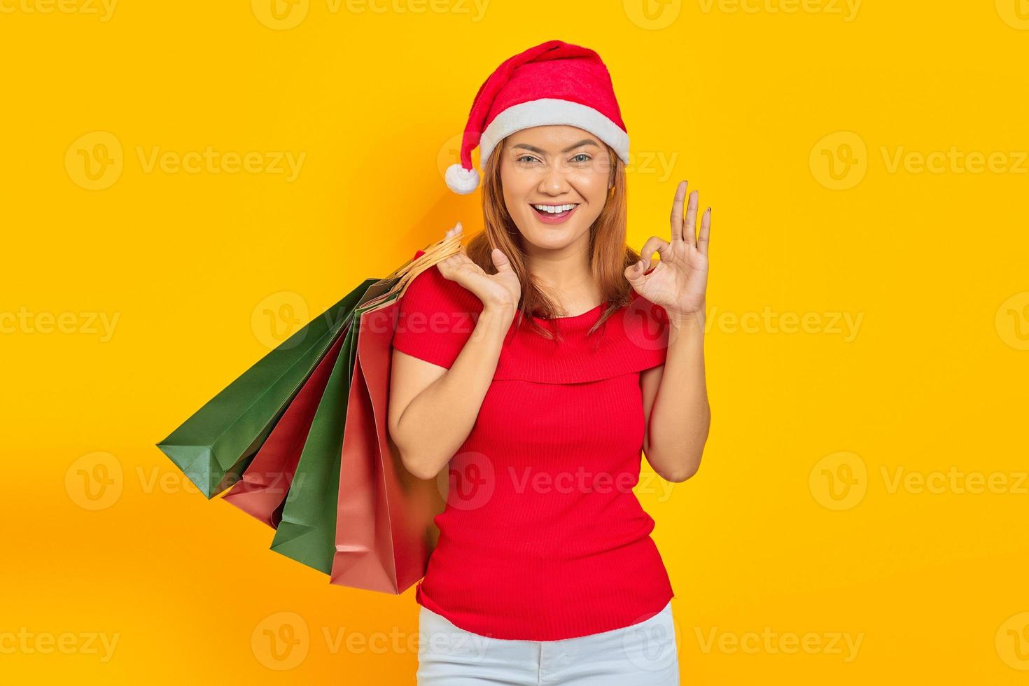 sorridente jovem mulher asiática com chapéu de Papai Noel segurando sacolas de compras e gesticulando um sinal de ok isolado sobre fundo amarelo foto