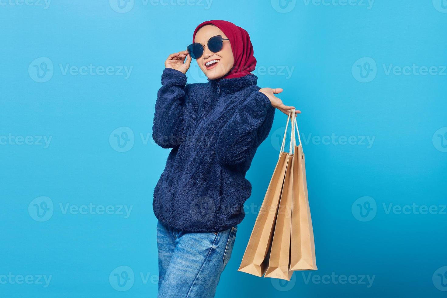 alegre jovem asiática usando óculos escuros e segurando sacolas de compras sobre fundo azul foto