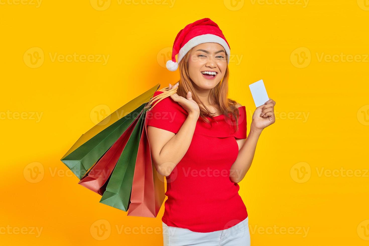 animada jovem asiática com chapéu de Papai Noel segurando sacolas de compras e um cartão de crédito em fundo amarelo foto