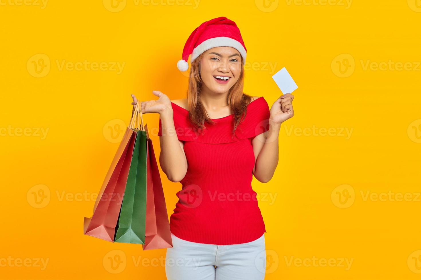 alegre jovem asiática com chapéu de Papai Noel segurando sacolas de compras e mostrando o cartão de crédito sobre fundo amarelo foto