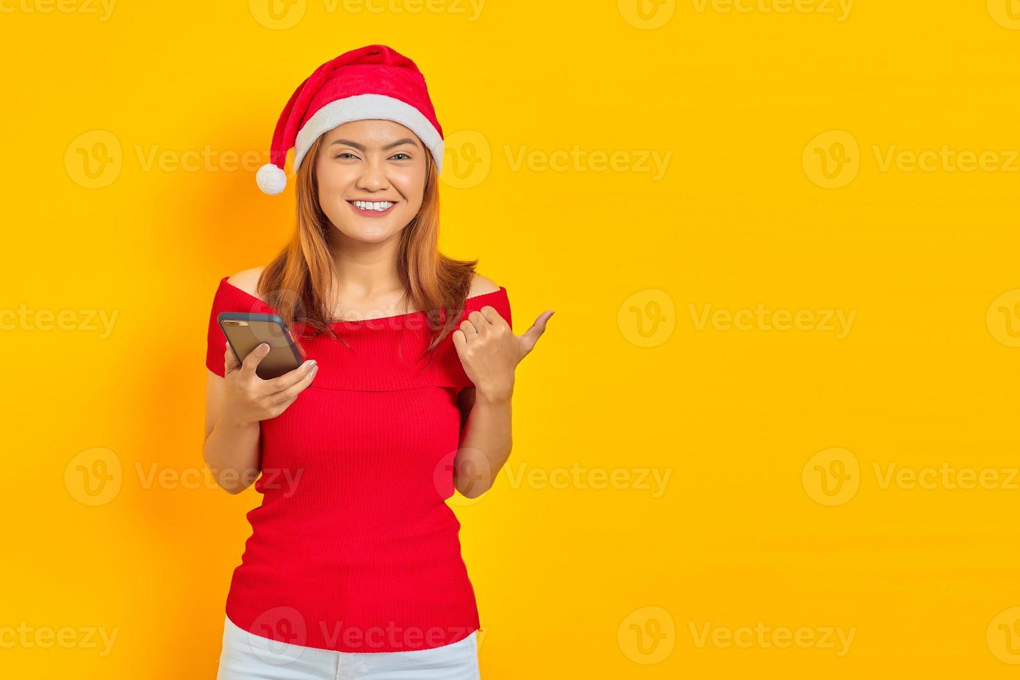 alegre jovem asiática com chapéu de Papai Noel segurando um telefone celular e mostrando os polegares isolados em um fundo amarelo foto
