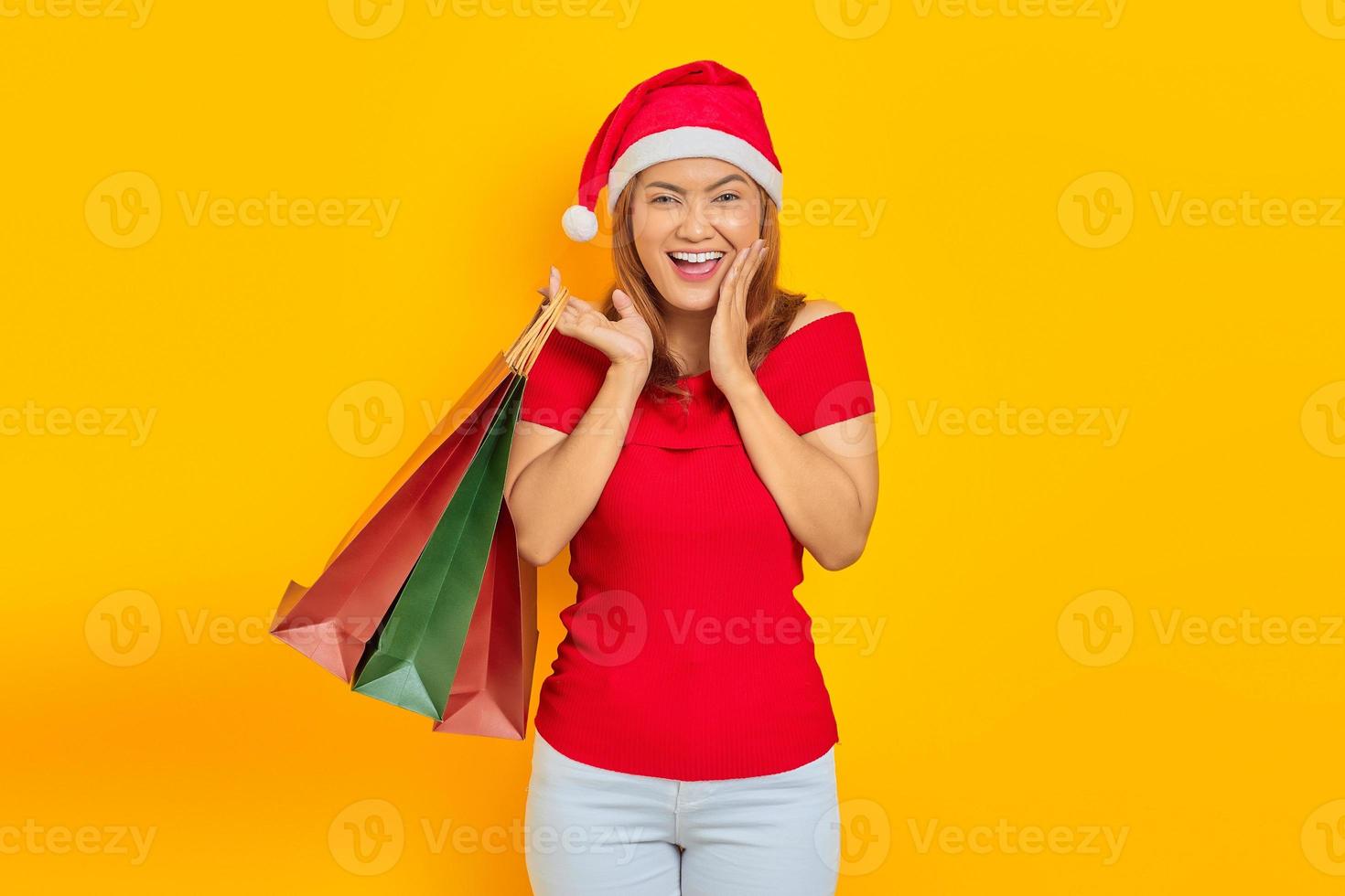 animada jovem asiática com chapéu de Papai Noel segurando sacolas de compras com a boca aberta sobre fundo amarelo foto