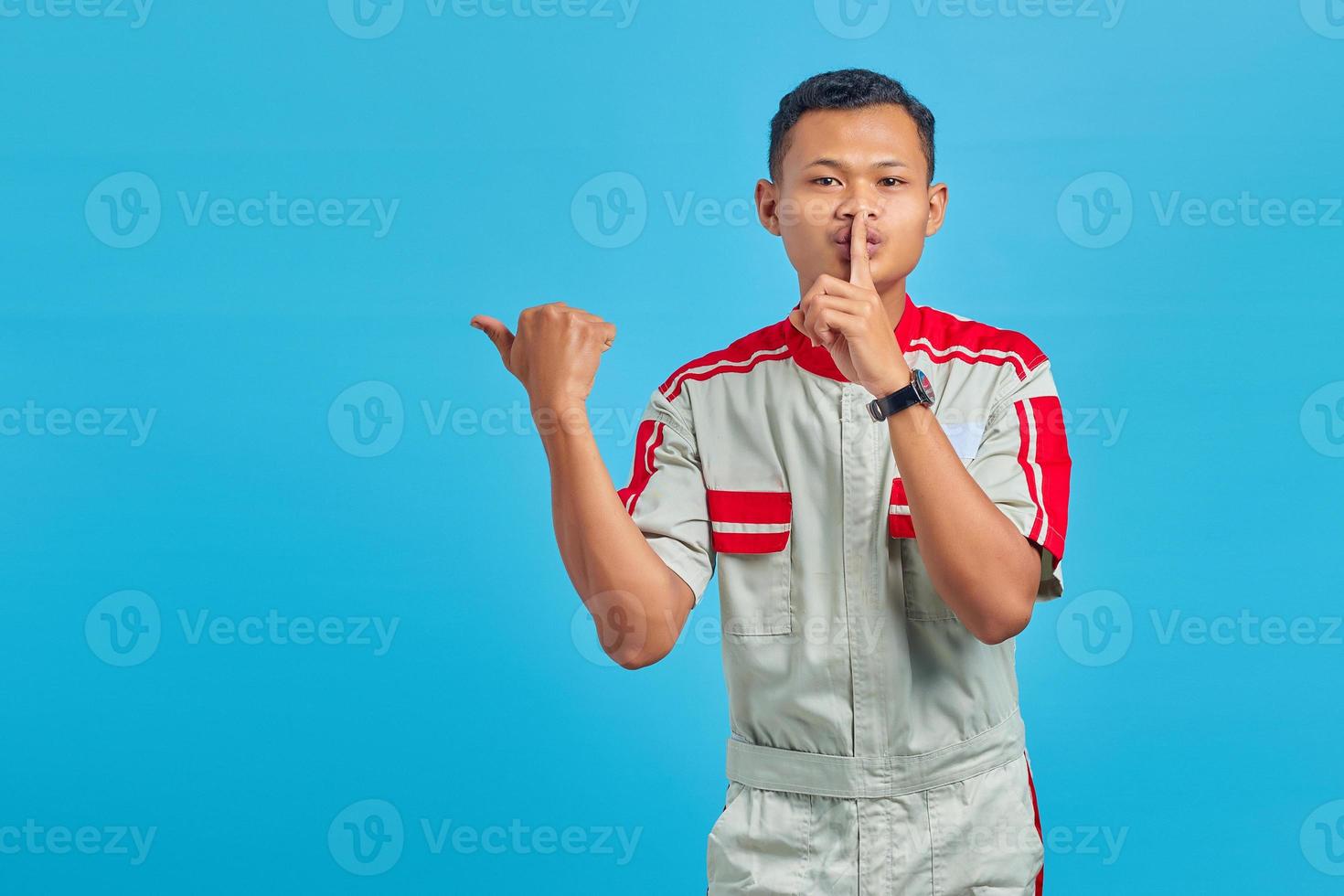 retrato de um jovem mecânico asiático sinalizando para ficar quieto e apontando para o lado sobre fundo azul foto