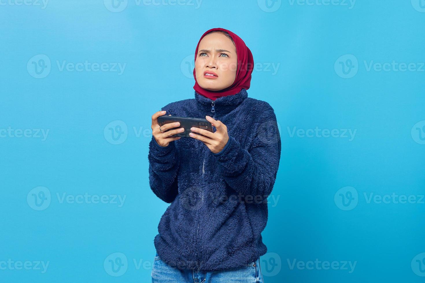 jovem asiática chocada jogando um videogame em um smartphone e olhando para cima sobre um fundo azul foto
