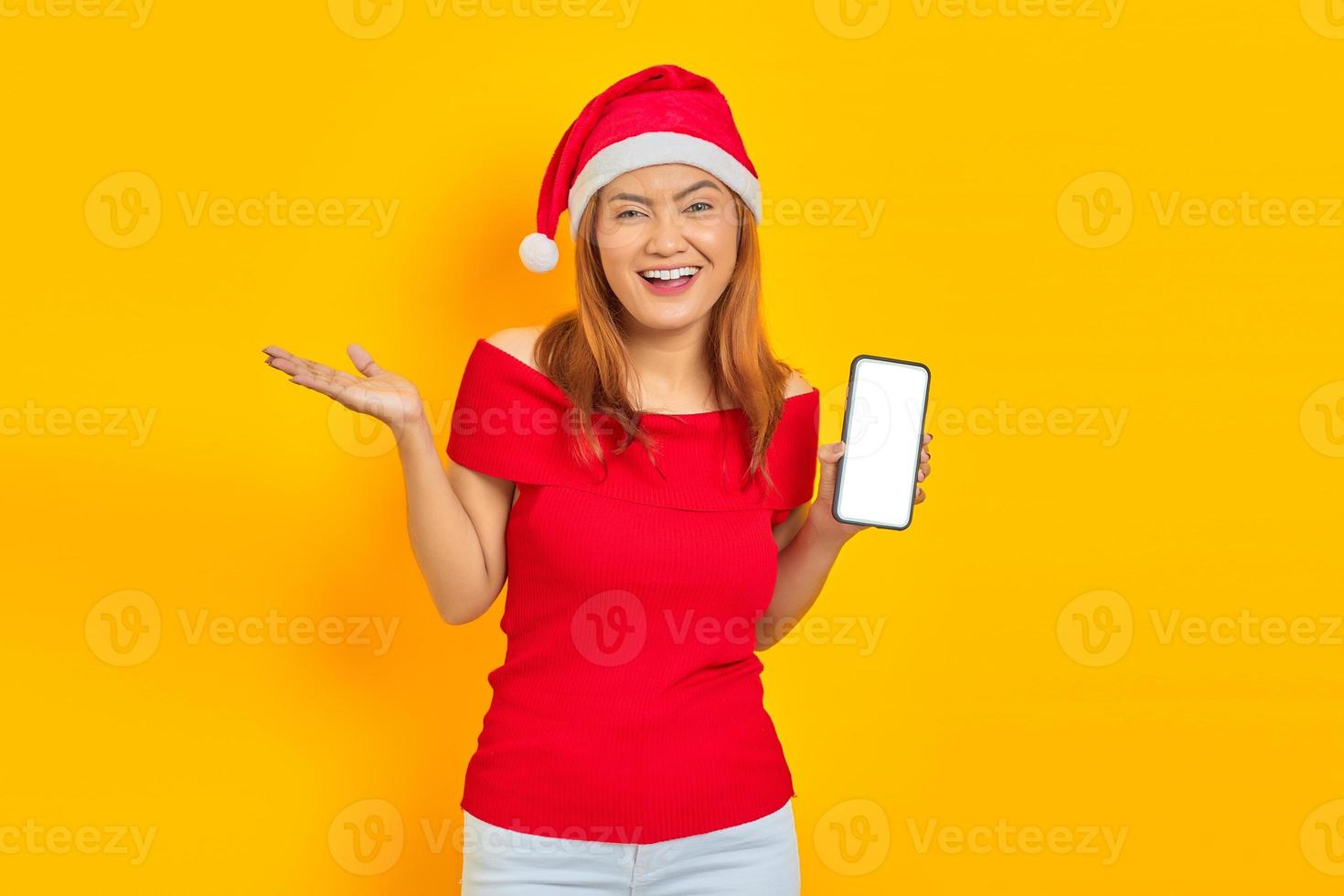 Mulher asiática jovem sorridente com chapéu de Papai Noel, estendendo a mão convidando para vir e mostrando o smartphone sobre fundo amarelo foto