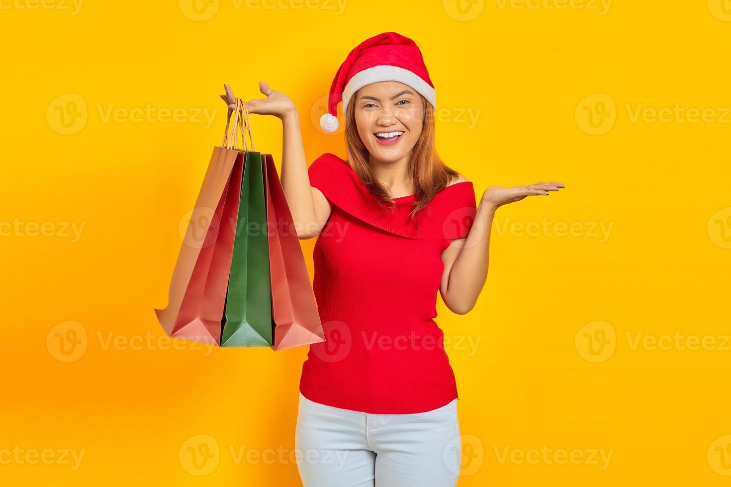 alegre jovem asiática com chapéu de Papai Noel segurando sacolas de compras e mostrando o espaço da cópia nas palmas das mãos sobre fundo amarelo foto