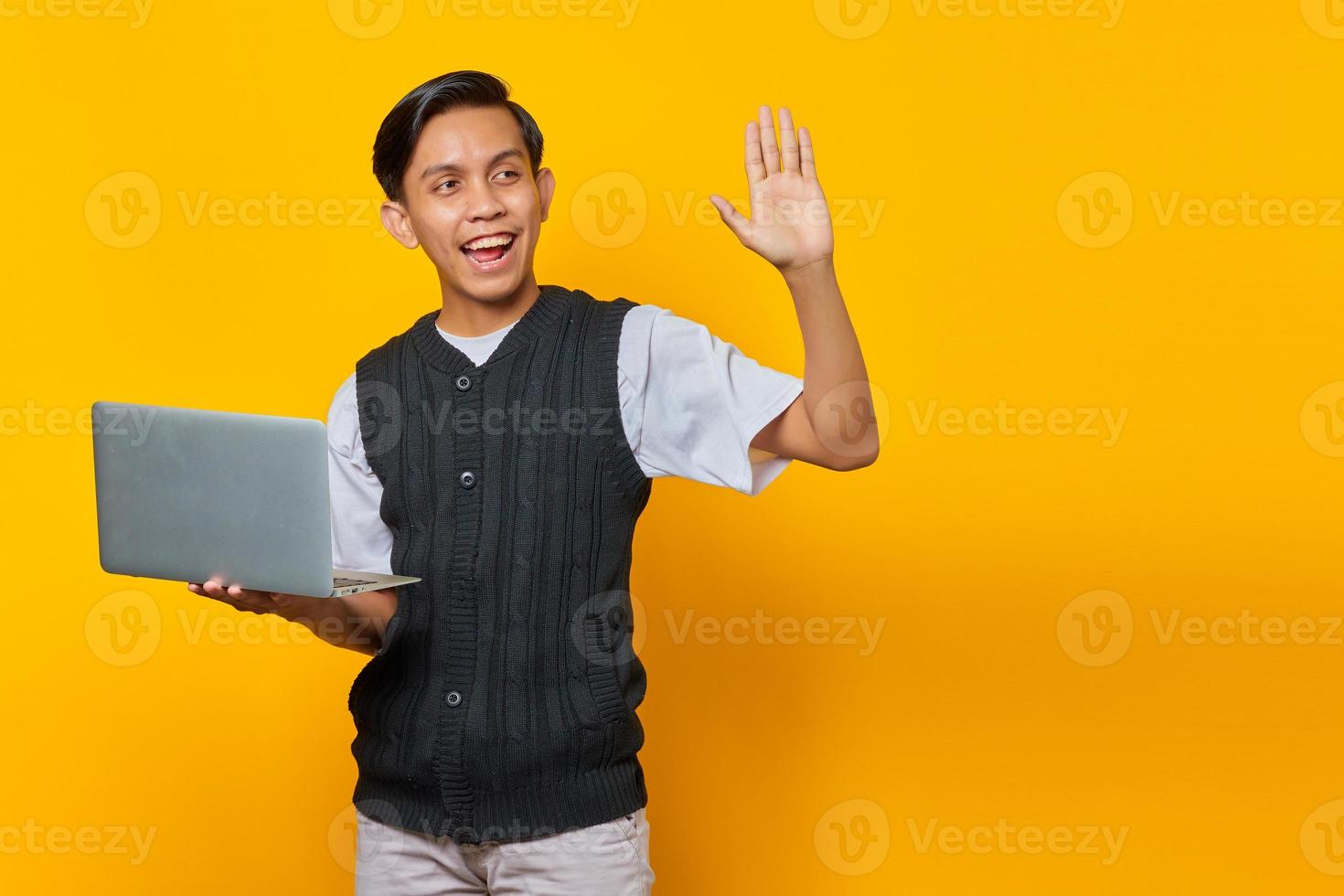 retrato de um jovem asiático sorridente segurando um laptop e gesticulando com a mão da onda sobre fundo amarelo foto