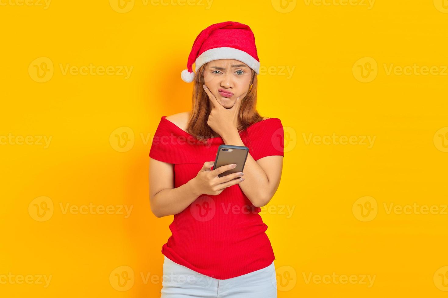 retrato de uma jovem mulher asiática com chapéu de Natal pensando e enviando mensagem em smartphone isolado em fundo amarelo foto