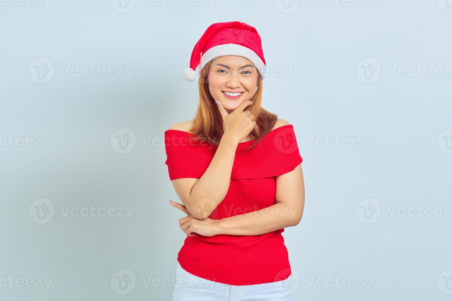 Mulher jovem e bonita com vestido de natal, sorrindo, olhando para a câmera com a mão tocando o queixo, isolado no fundo branco foto