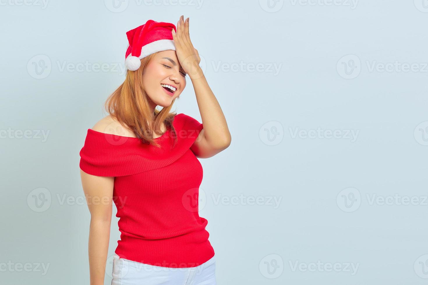 Surpreendeu a jovem mulher asiática com chapéu de Natal e pressionando as mãos na cabeça em fundo branco foto