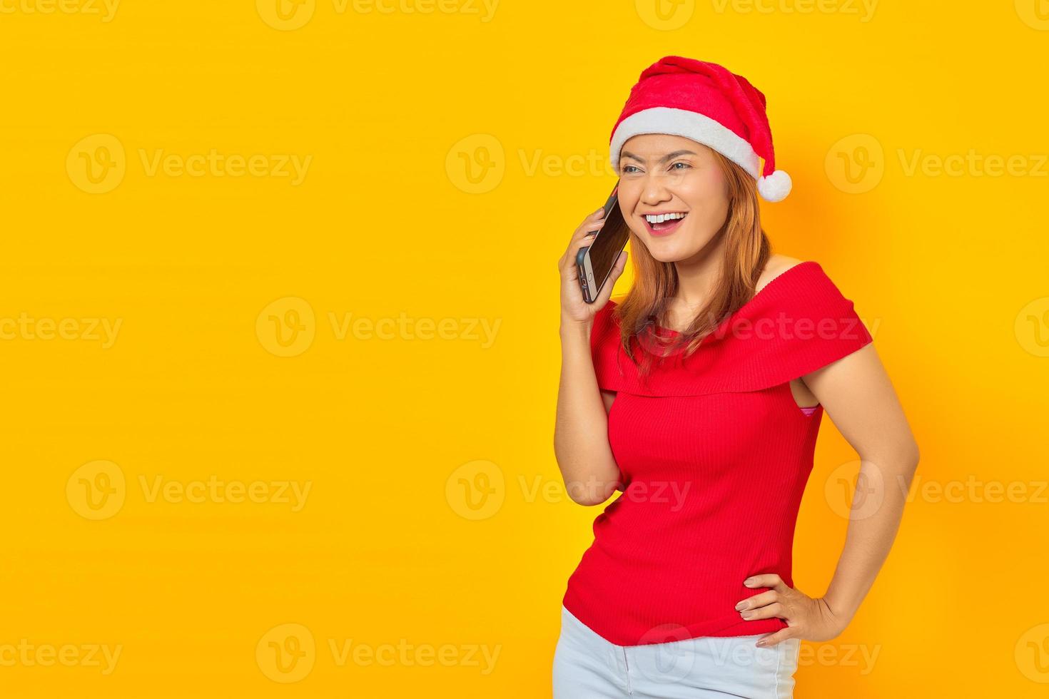 alegre jovem asiática usando chapéu de natal falando no smartphone e colocando as mãos na cintura em fundo amarelo foto